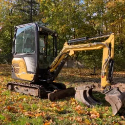 Żółto-czarna mini koparka na gąsienicach stoi na trawie pokrytej jesiennymi liśćmi w Zgierzu. Dwie dodatkowe łyżki obok. W tle drzewa z żółknącymi liśćmi.