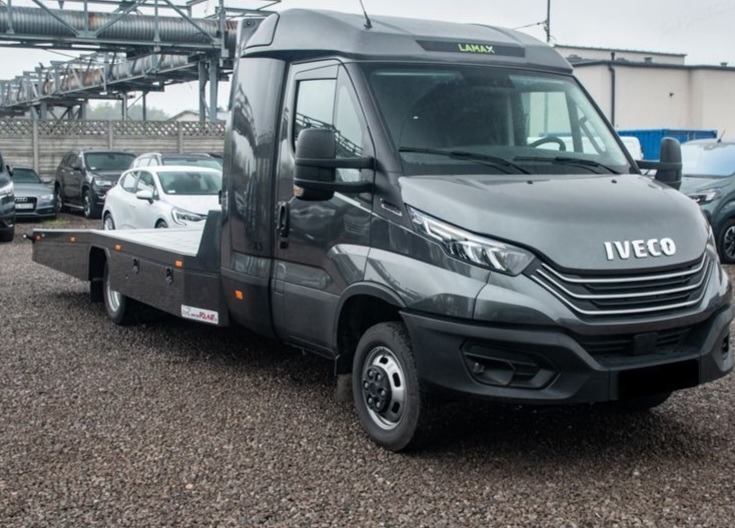 Szara laweta Iveco na żwirowym placu, widok z boku, z metalową konstrukcją w tle. Widoczne samochody osobowe w oddali.