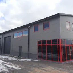 Nowoczesna hala przemysłowa z szarych płyt warstwowych z czerwonymi ramami okiennymi i drzwiowymi, częściowo pokryta resztkami śniegu.