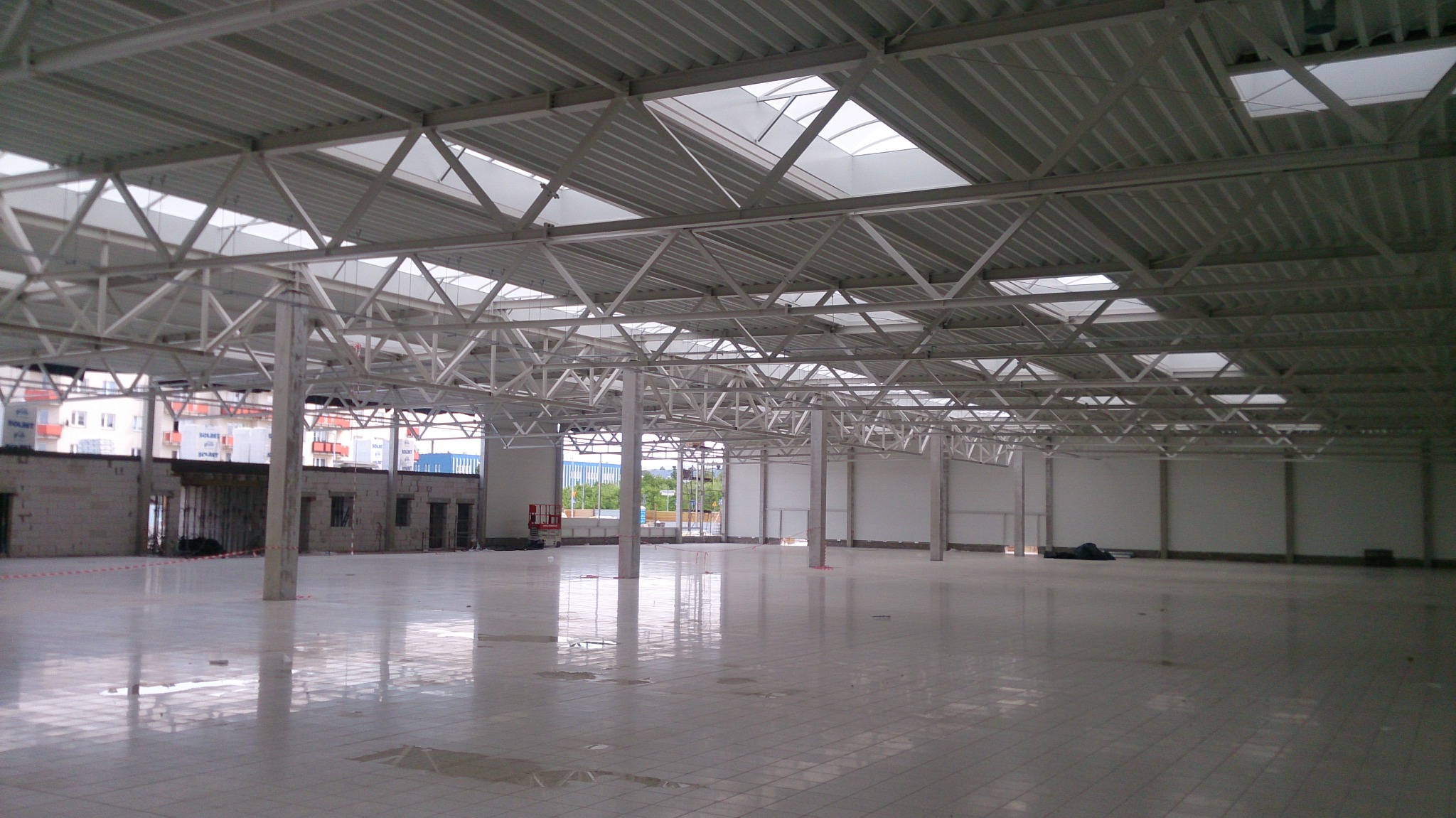 Wnętrze hali w trakcie budowy z widoczną konstrukcją stalową dachu i słupami nośnymi, surowa posadzka z widocznymi plamami wody.