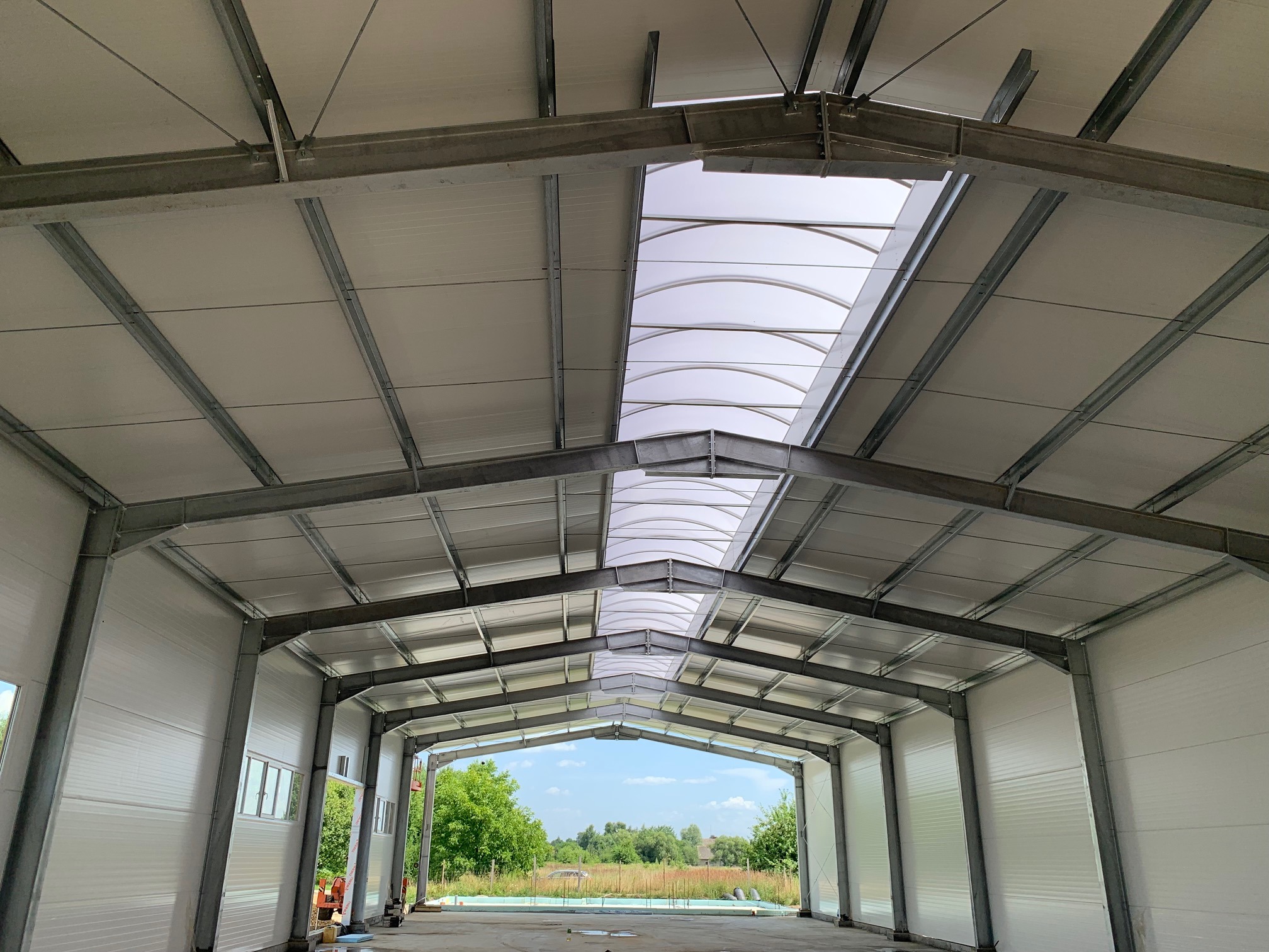 Wnętrze hali w trakcie budowy, widoczne stalowe konstrukcje nośne, białe płyty warstwowe na ścianach i dachu, oraz świetliki dachowe w kształcie łuków, z widokiem na zewnątrz przez otwarty wjazd.