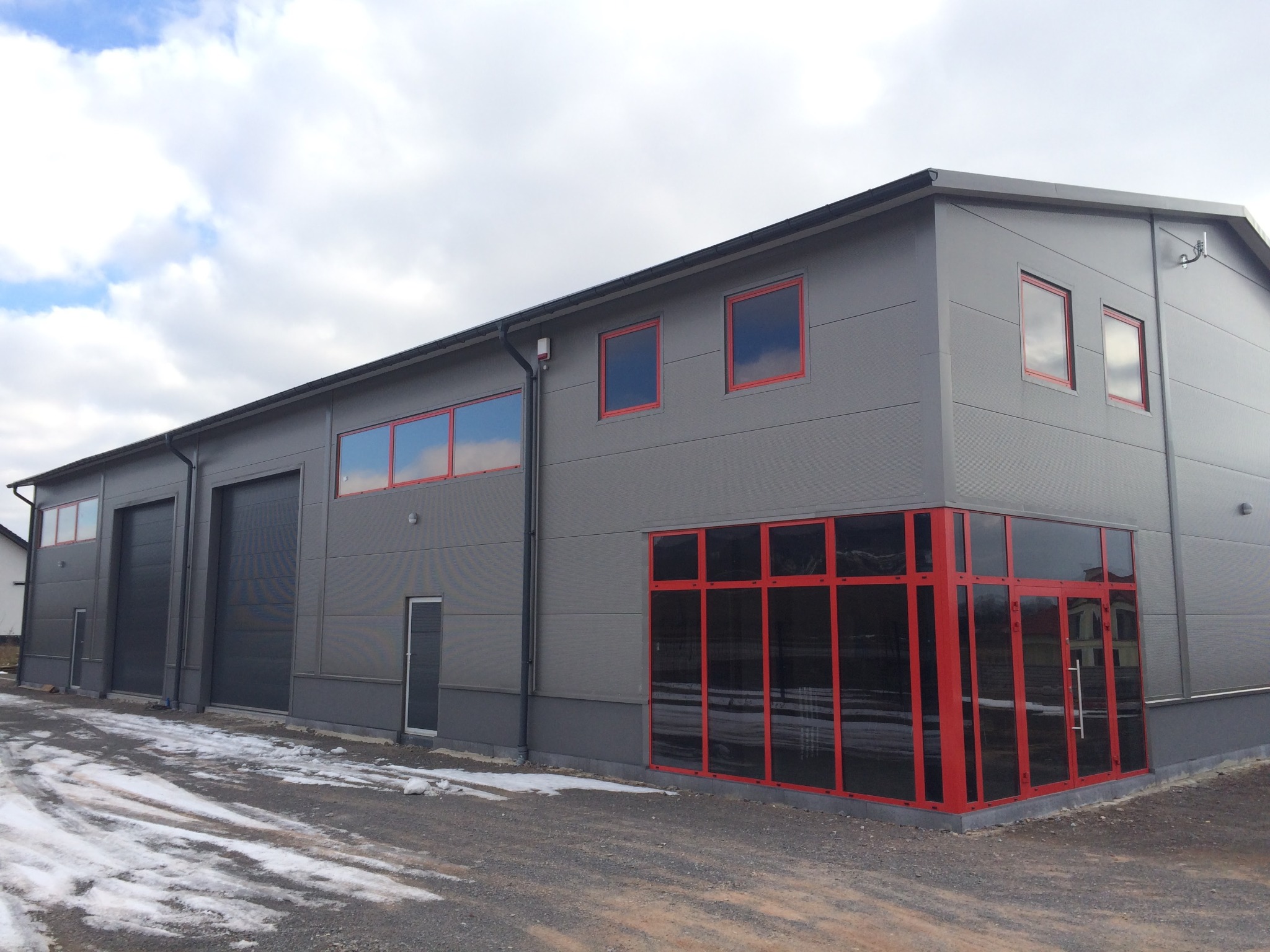 Nowoczesna hala przemysłowa z szarych płyt warstwowych z czerwonymi ramami okiennymi i drzwiowymi, częściowo pokryta resztkami śniegu.