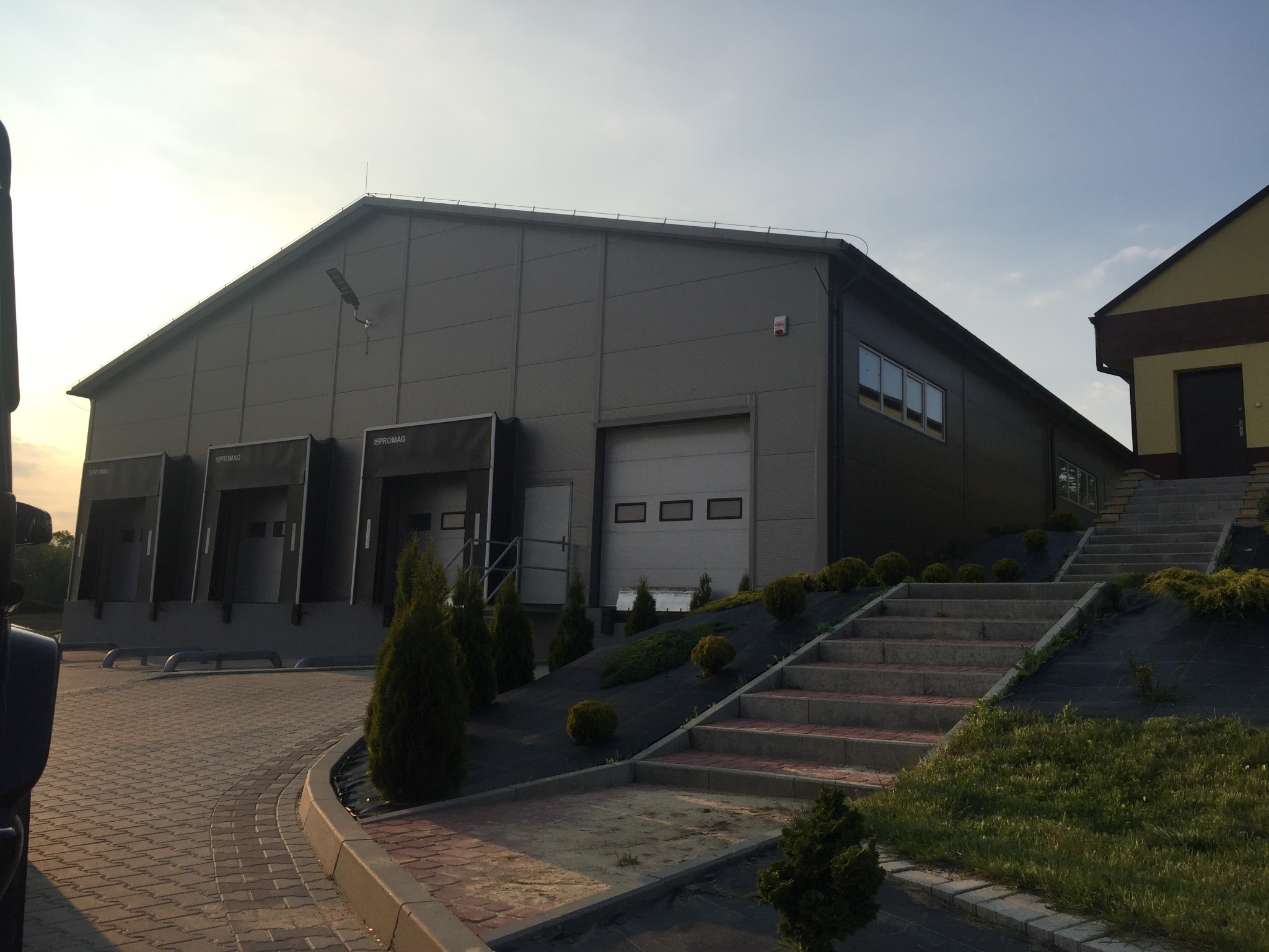 Nowoczesna hala magazynowa z rampami załadunkowymi w Starachowicach, widok z zewnątrz, z kostką brukową i schodami prowadzącymi do budynku biurowego.