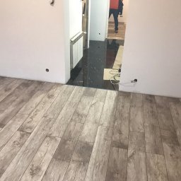 Ich bud - Ułożona podłoga z paneli imitujących drewno w nowym mieszkaniu, widoczne gniazdka elektryczne w ścianie i fragment czarnej, błyszczącej podłogi w korytarzu z grzejnikiem.