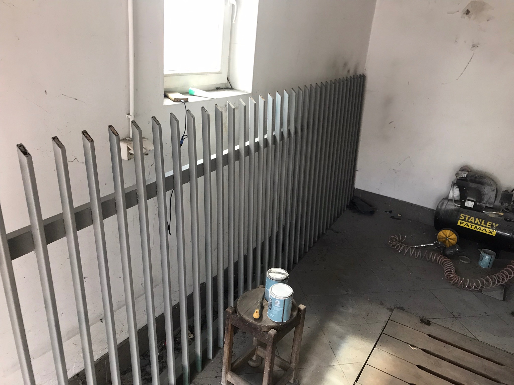 Malowanie grzejnika żeberkowego na szaro, widoczne puszki z farbą na stołku i kompresor Stanley Fatmax w tle, surowy stan pomieszczenia.