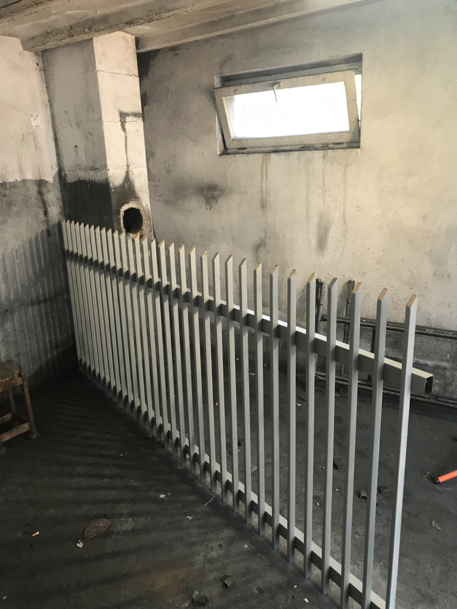 Szara, metalowa balustrada ochronna, częściowo zamontowana w surowym pomieszczeniu z betonowymi ścianami i małym oknem.