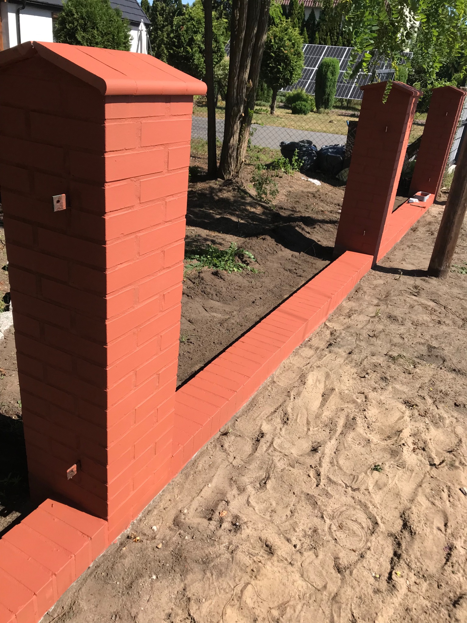 Czerwona, pomalowana ceglana podmurówka ogrodzenia w trakcie budowy, widoczne słupki i fragmenty muru, ziemia wokół, w tle dom i panele słoneczne.