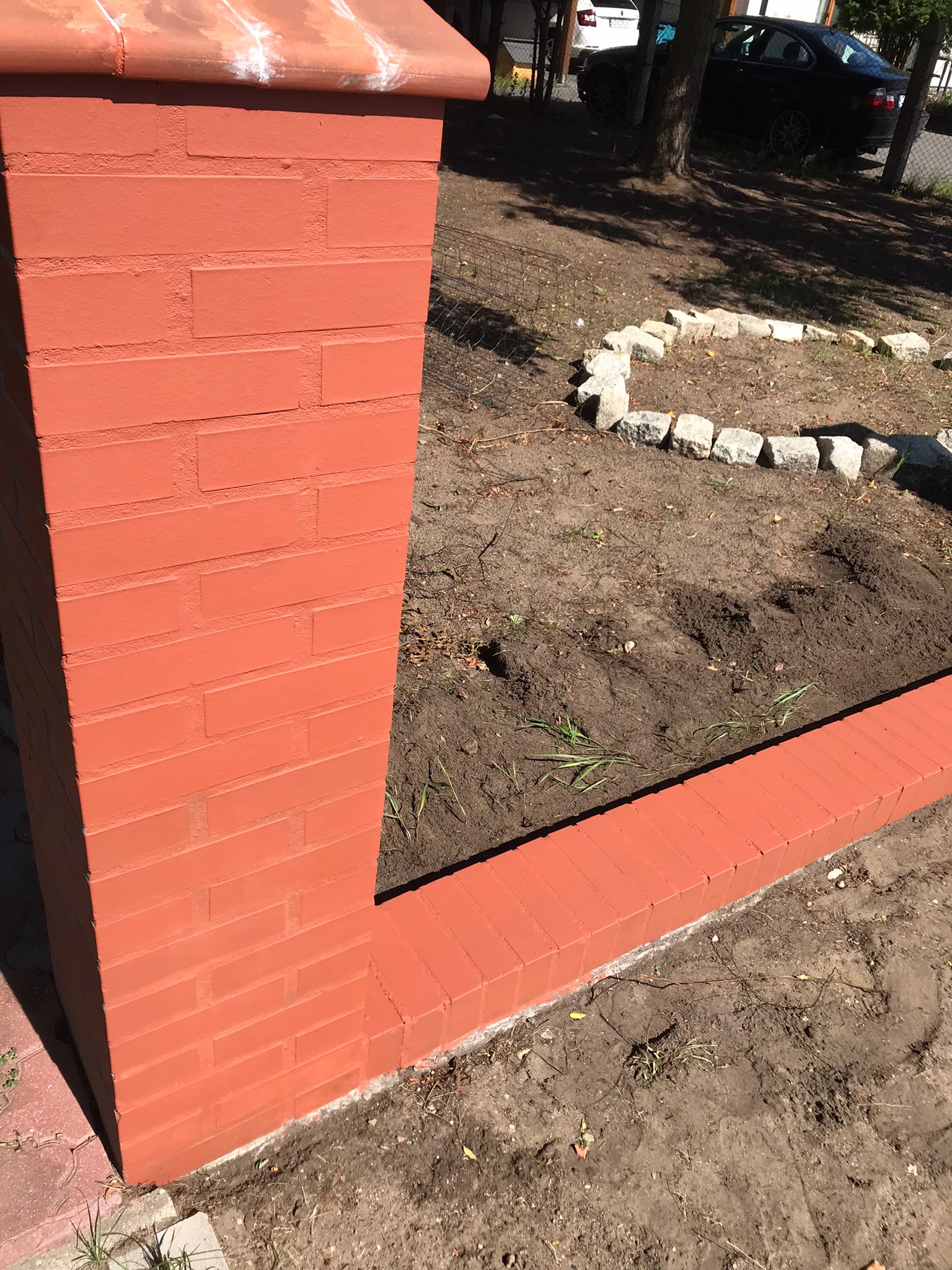 Pomarańczowy, pomalowany ceglany słupek ogrodzeniowy z fragmentem niskiego murku otaczającego rabatę, otoczenie w trakcie prac ziemnych.