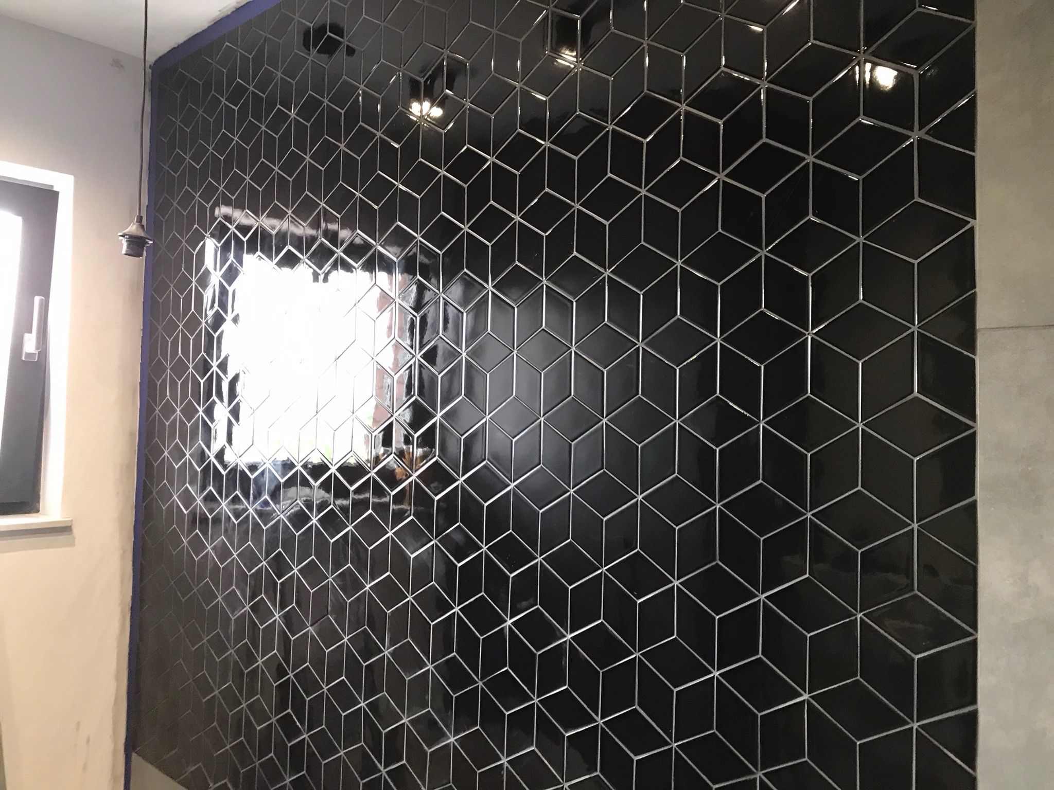Ściana wyłożona błyszczącymi czarnymi płytkami w geometryczny wzór 3D, odbijające światło, z fragmentem okna i lampy po lewej stronie.