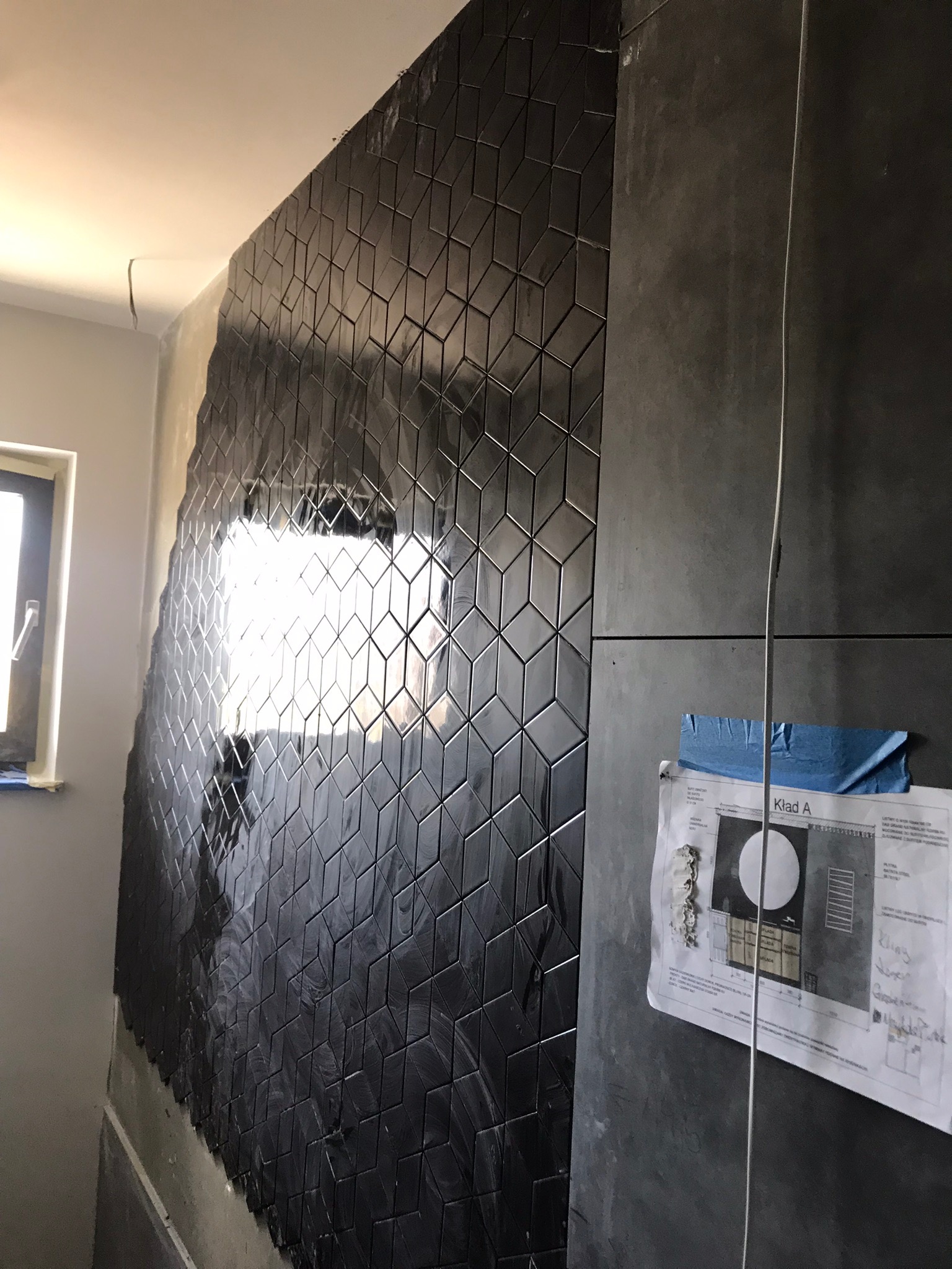 Ściana łazienki w trakcie wykańczania z błyszczącą, czarną mozaiką o geometrycznym wzorze 3D, obok ściana z dużych szarych płyt, na której przyklejony jest plan łazienki.