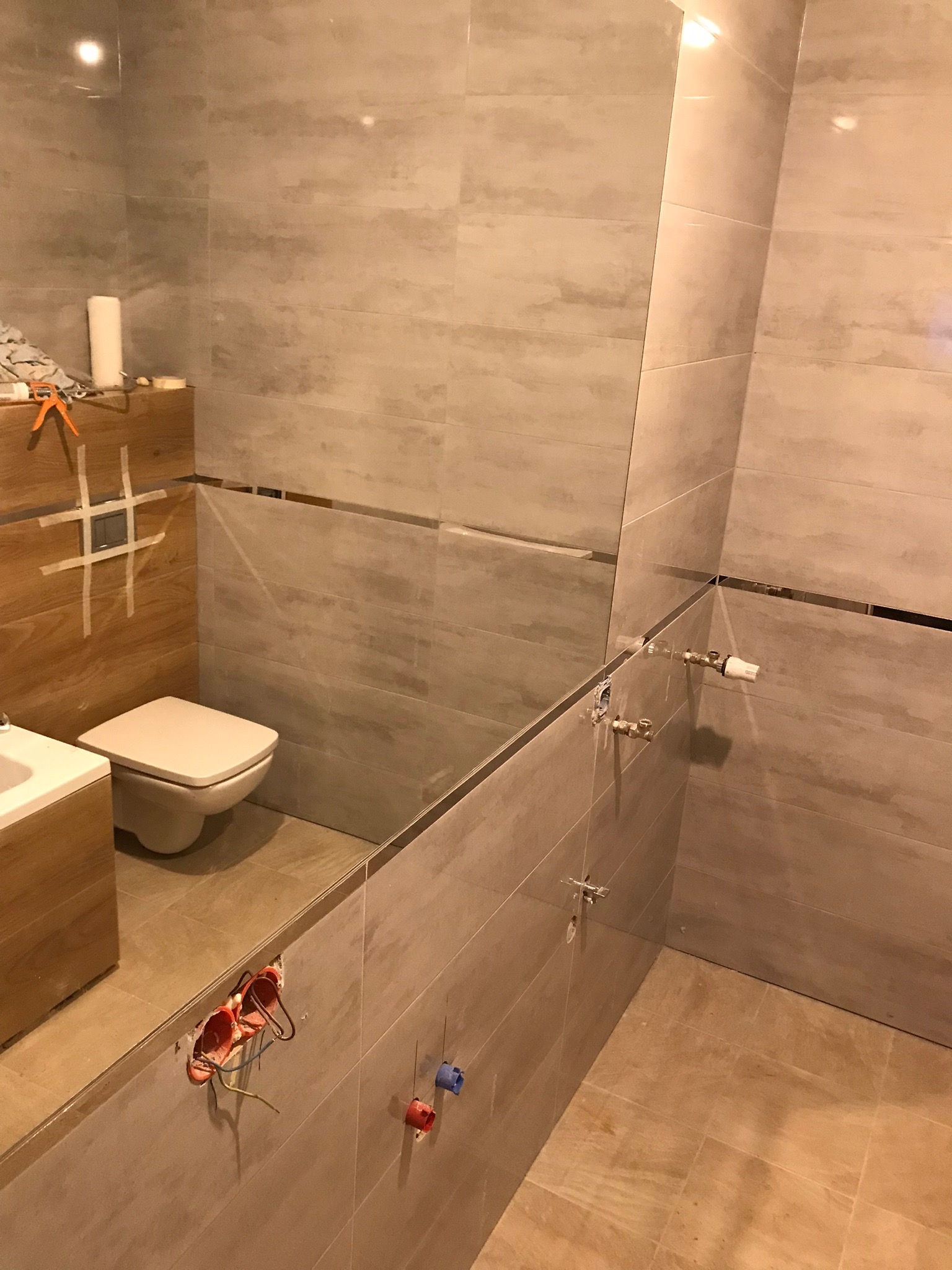 Wnętrze łazienki w trakcie remontu, widoczne płytki imitujące kamień na ścianach i podłodze, niezabudowane instalacje wodne i elektryczne, oraz toaleta z elementami maskowania taśmą malarską.