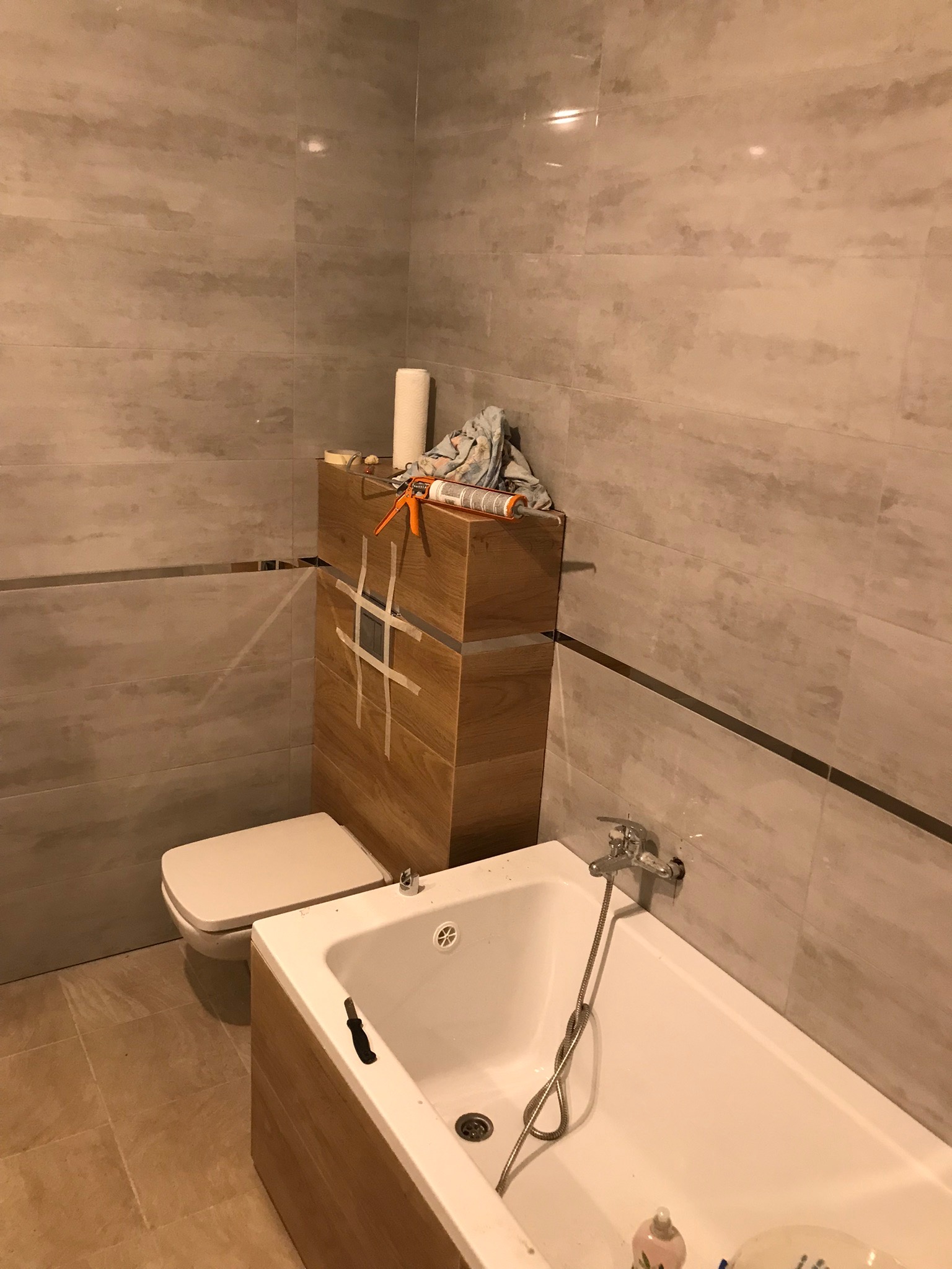 Łazienka w trakcie remontu: płytki imitujące marmur na ścianach, zabudowa WC z przyklejoną taśmą malarską, wanna z baterią, materiały i narzędzia na szafce.