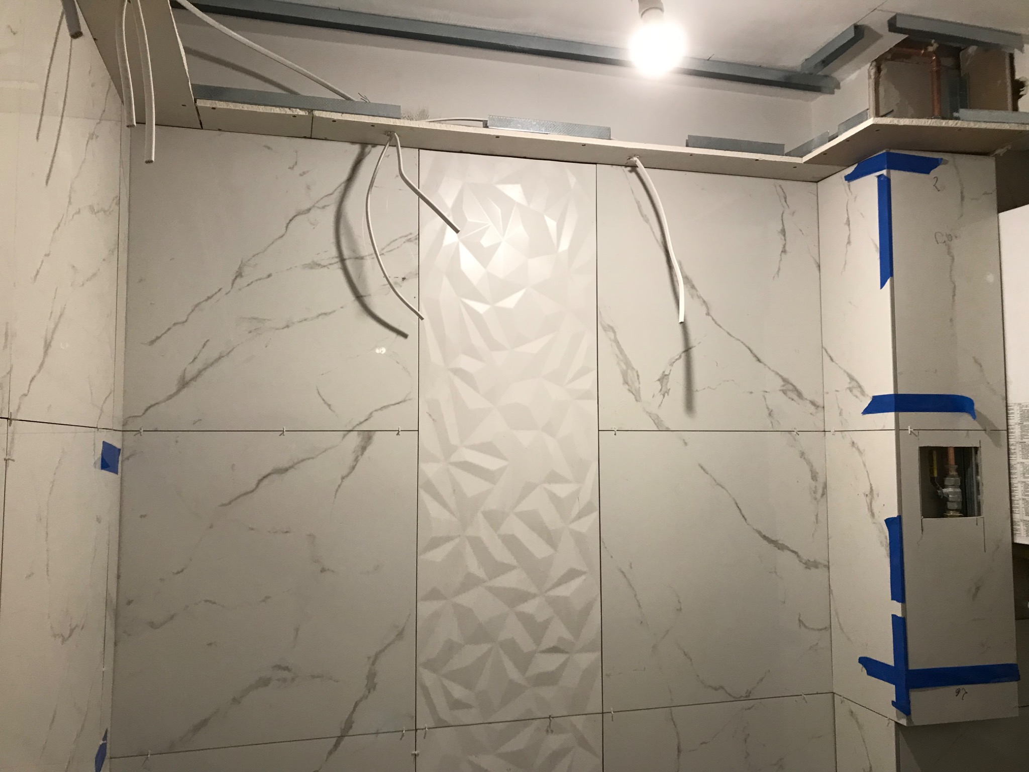 Ściana łazienki w trakcie układania płytek: białe płytki z szarym marmurkowym wzorem, w tym jedna pionowa sekcja z trójwymiarowym wzorem geometrycznym, widoczne niepodłączone kable elektryczne...