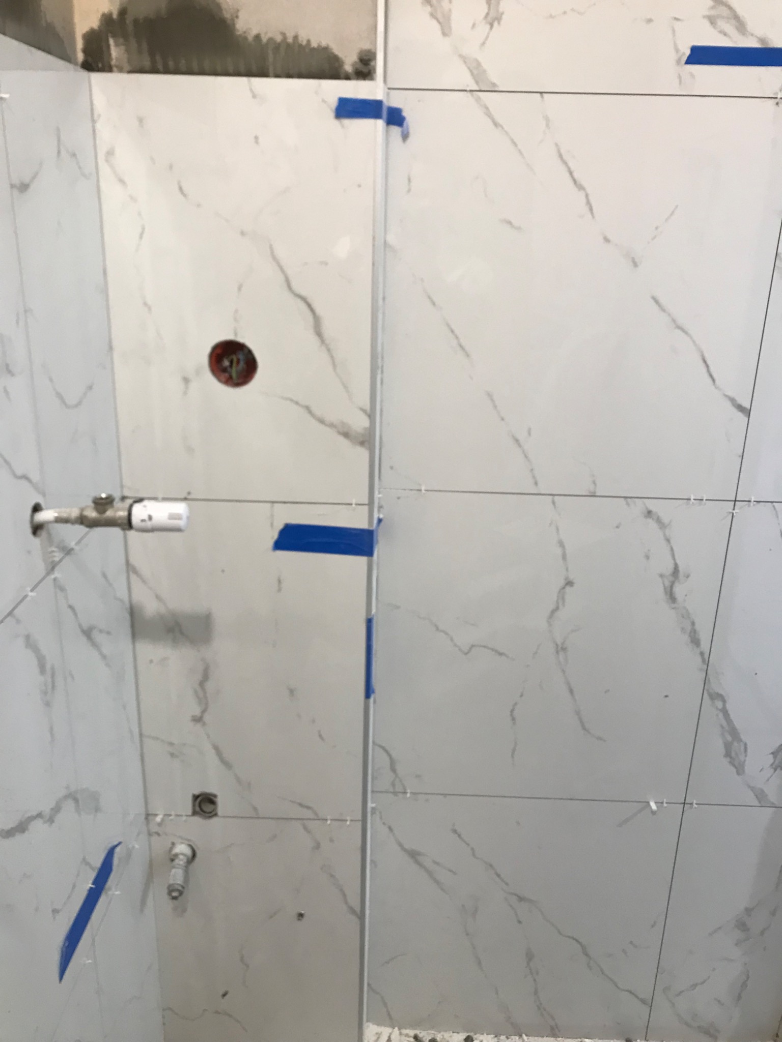 Fragment ściany w trakcie układania białych płytek imitujących marmur, z widocznymi niebieskimi taśmami mocującymi i krzyżykami dystansowymi, oraz wystającymi rurami i puszką elektryczną.