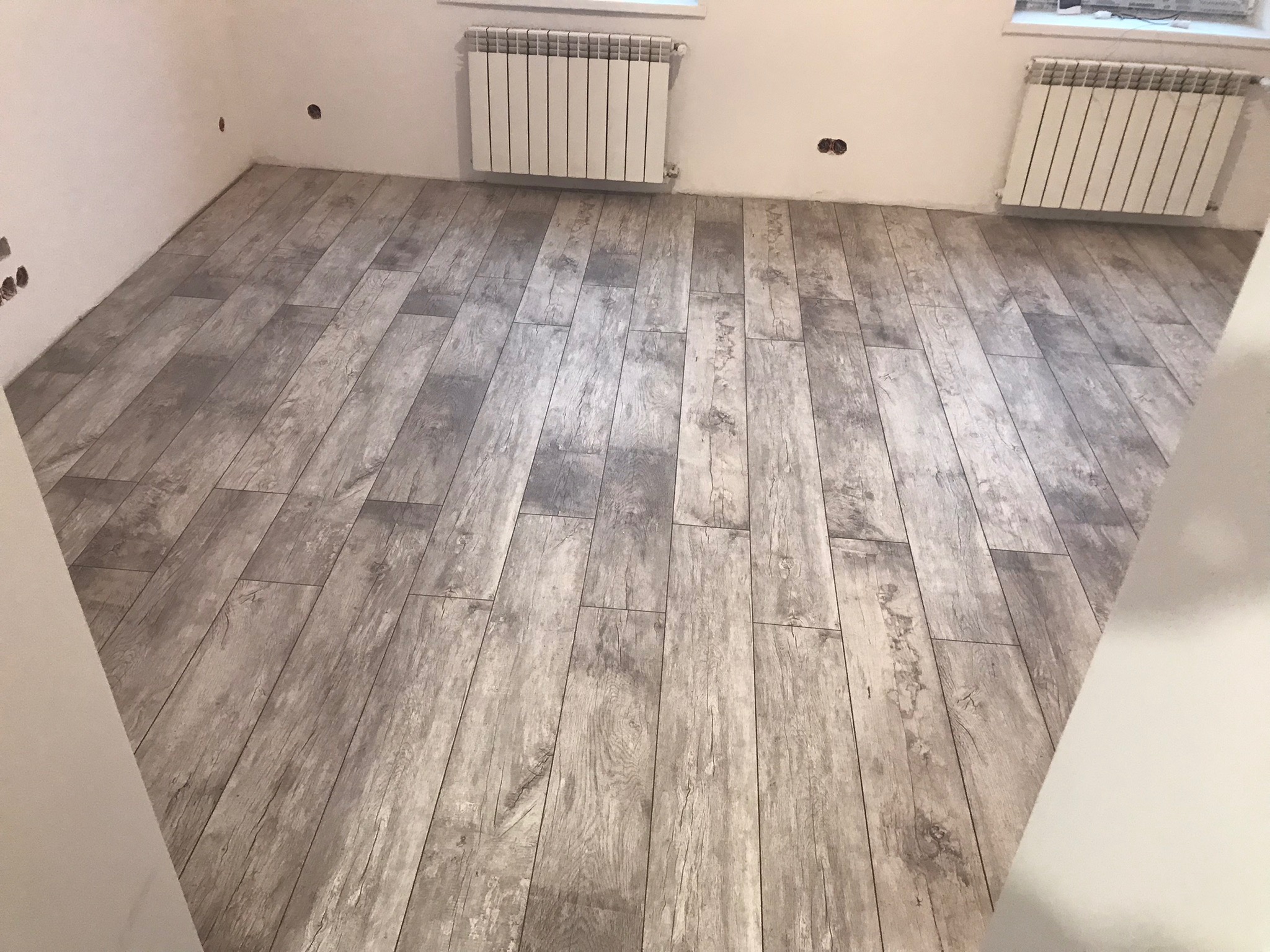 Ułożona podłoga z paneli imitujących drewno w odcieniach szarości, widoczne grzejniki na ścianie i puszki elektryczne bez osprzętu.