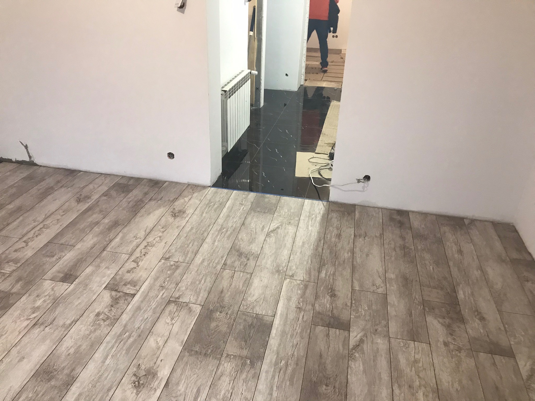 Ułożona podłoga z paneli imitujących drewno w nowym mieszkaniu, widoczne gniazdka elektryczne w ścianie i fragment czarnej, błyszczącej podłogi w korytarzu z grzejnikiem.