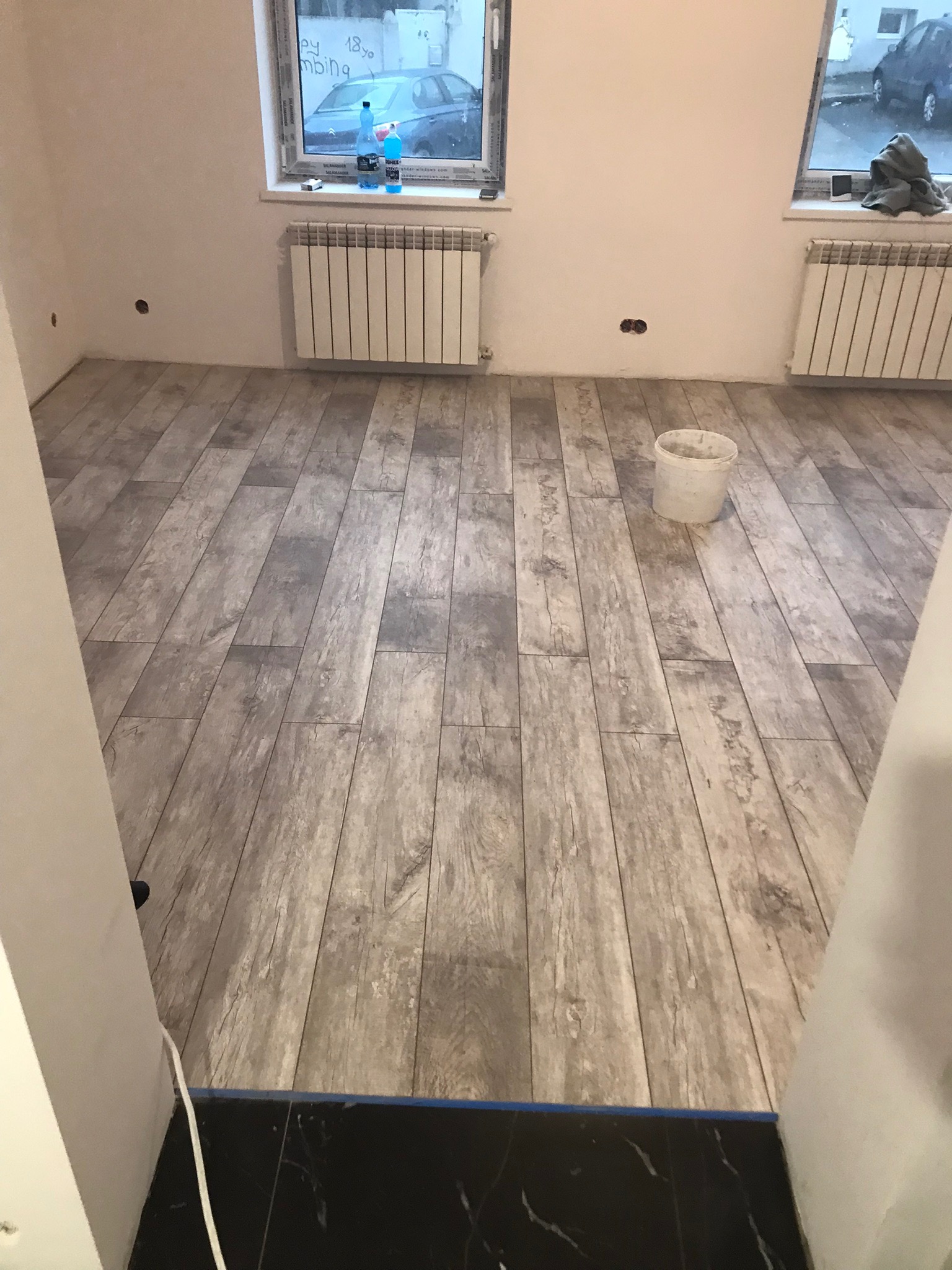 Ułożona podłoga z płytek imitujących drewno w odcieniach szarości, widoczne grzejniki pod oknami, białe ściany z otworami na gniazdka i wiadro po farbie.