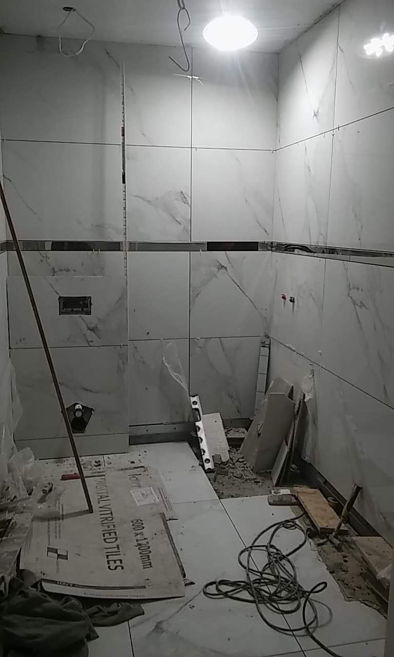 Wnętrze łazienki w trakcie prac wykończeniowych, widoczne białe płytki imitujące marmur na ścianach, częściowo ułożone, narzędzia glazurnicze, karton po płytkach na podłodze, widoczne fragmenty...