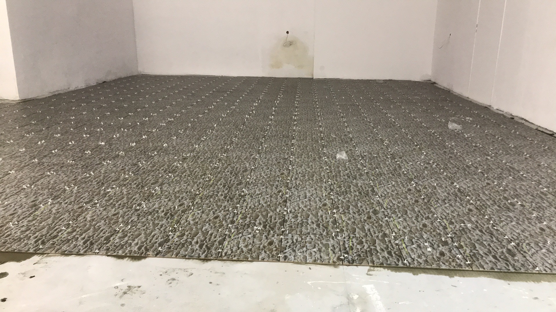 Szare płytki podłogowe ułożone w nowym pomieszczeniu, z białymi krzyżykami dystansowymi, widoczne ślady zaprawy na podłodze.