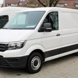 Biały, długi furgon Volkswagen Crafter na kostce brukowej, widok z boku, lekko z przodu. Drugi, podobny furgon widoczny w tle, po lewej stronie.