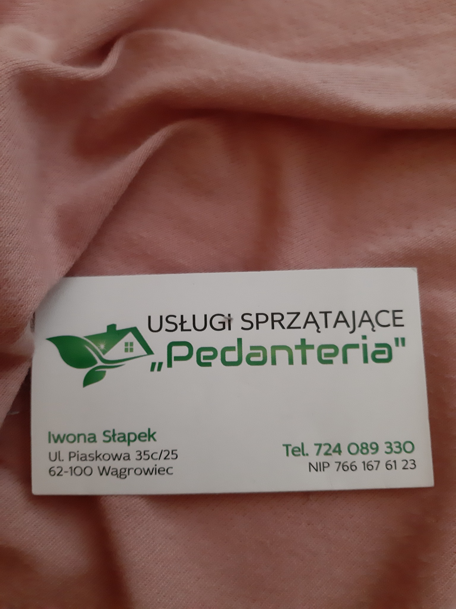 Wizytówka firmy sprzątającej 'Pedanteria' na tle lekko pogniecionej, różowej tkaniny. Wizytówka zawiera logo z zielonym liściem i zarysem domu, dane kontaktowe Iwony Stapek, adres w Wągrowcu...