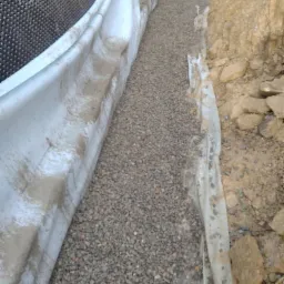 Wykonanie drenażu opaskowego fundamentów z użyciem folii kubełkowej i warstwy żwiru, widoczne wykopane podłoże.