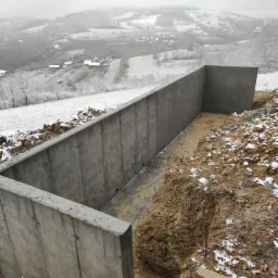 Surowy betonowy mur oporowy w kształcie litery L, w wykopie ziemnym, z widokiem na zasypany śniegiem krajobraz wiejski w tle, pochmurny dzień.