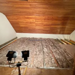 Ocieplenie poddasza wełną mineralną, widoczne drewniane belki, oświetlenie robocze na statywie i drewniana boazeria na suficie.