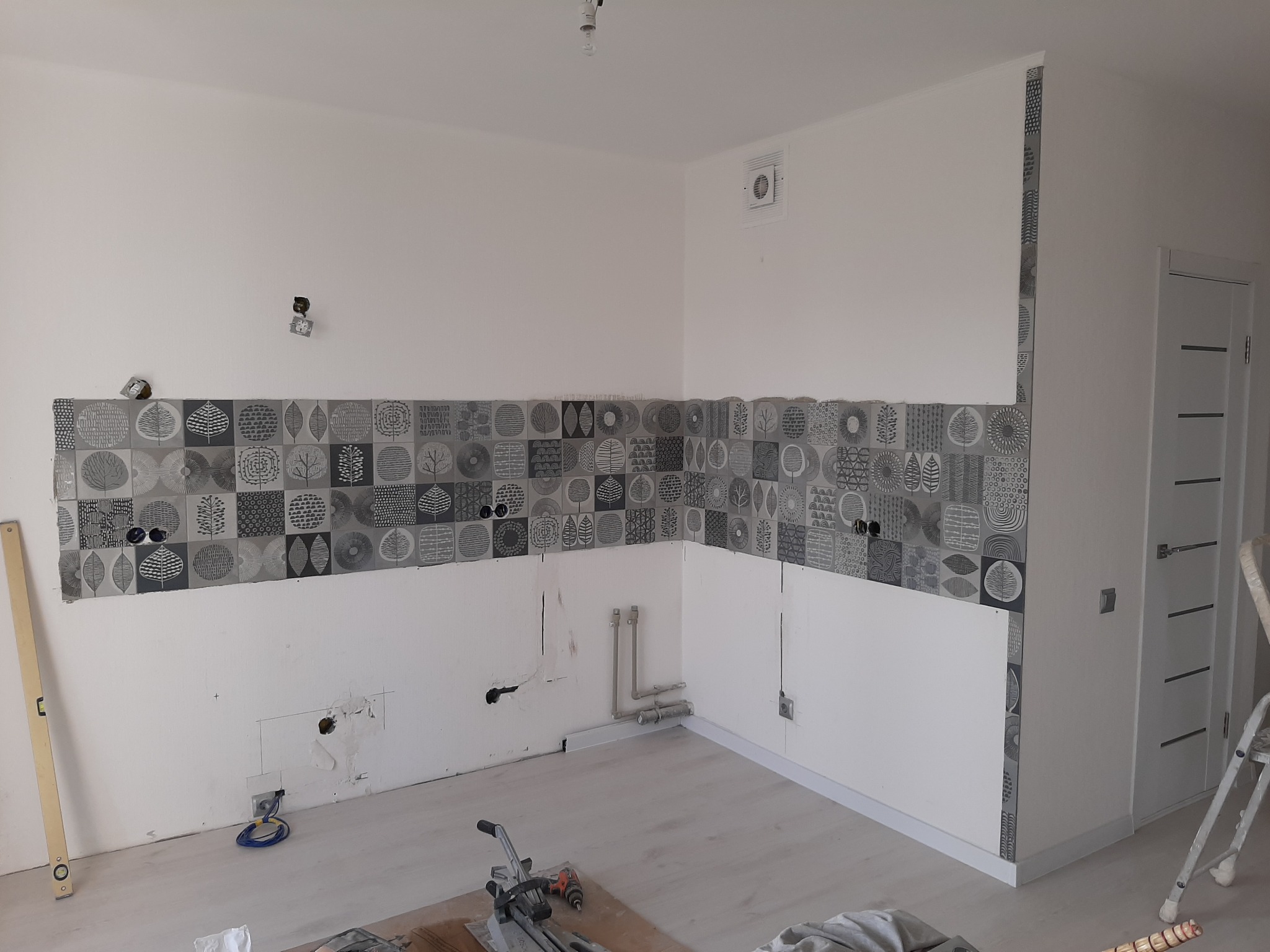 Fragment ściany w trakcie układania dekoracyjnych płytek ceramicznych z motywem drzew na wysokości wzroku, widoczne przygotowane instalacje elektryczne i hydrauliczne, poziomica i maszyna do cięcia...