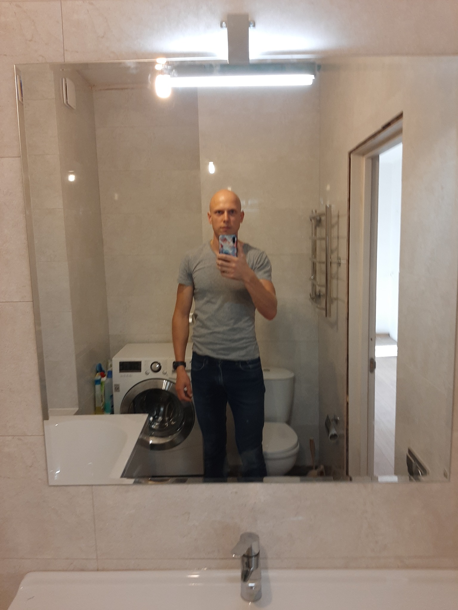 Odbicie mężczyzny w lustrze łazienki z pralką, toaletą i umywalką w tle, widoczne płytki ceramiczne na ścianach.
