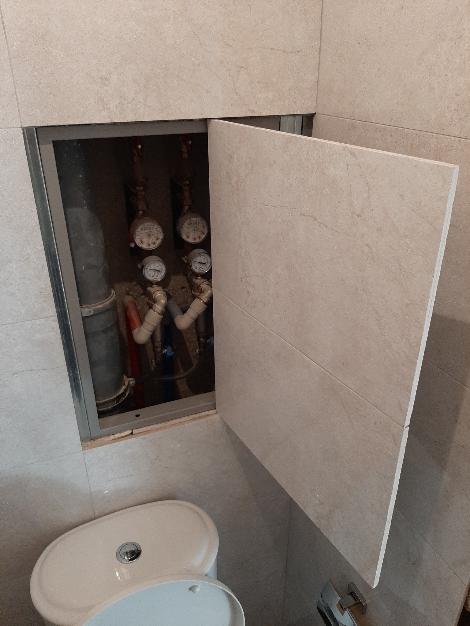 Otwarta rewizja wodomierzowa w łazience, ukryta za drzwiczkami z płytek ceramicznych, widoczne rury i liczniki.