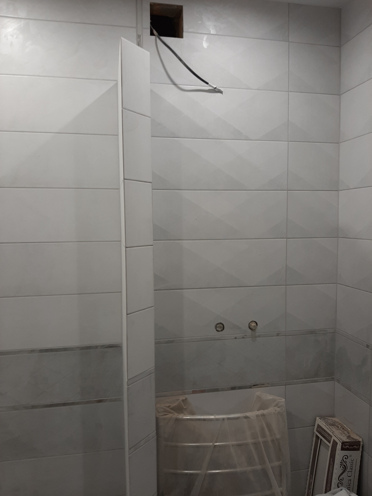 Łazienka w trakcie remontu: białe płytki z delikatnym wzorem, wystające kable i rury, zabezpieczona folią wanna, karton z płytkami w rogu.