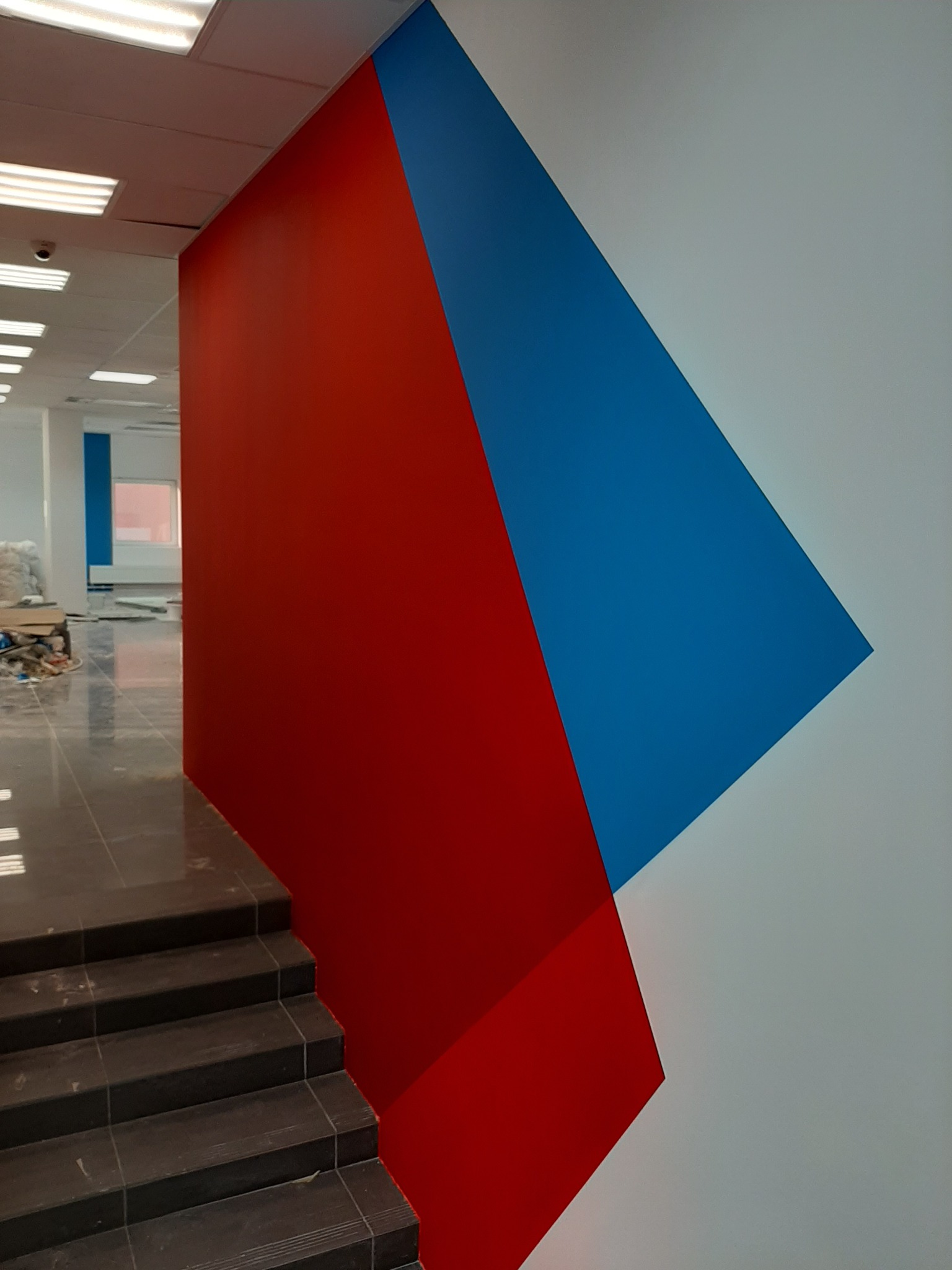 Ściana biura z geometrycznym malowaniem w kolorach czerwonym, niebieskim i białym, widoczne schody z ciemnego kamienia i fragment pomieszczenia w tle.