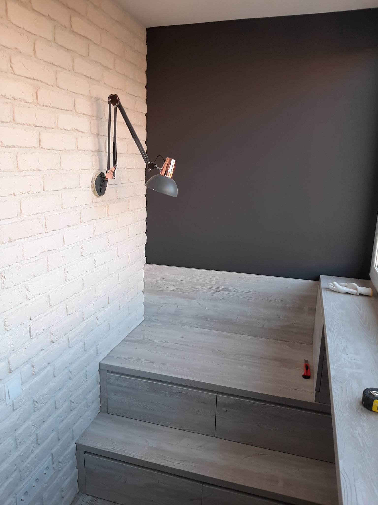 Biała ściana z cegły pomalowana na biało z czarną lampą na wysięgniku, obok ciemnoszara ściana i drewniane schody z szufladami, w tle okno i narzędzia.