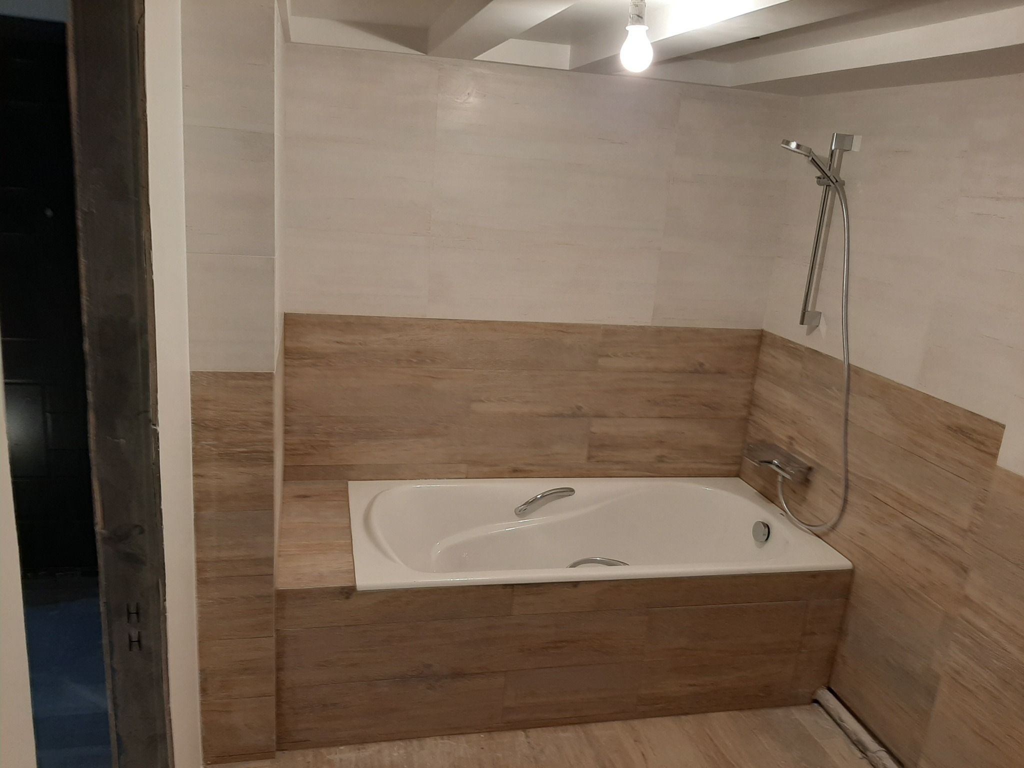 Wykończona łazienka z wanną obudowaną płytkami imitującymi drewno, jasne płytki na ścianach, widoczna słuchawka prysznicowa i fragment niedokończonego wejścia do pomieszczenia.