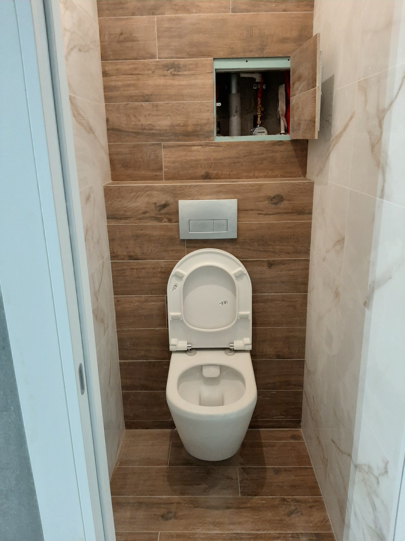 Nowoczesna toaleta z podwieszanym sedesem i ukrytą spłuczką, obłożona płytkami imitującymi drewno i marmur, z otwartą klapką rewizyjną nad sedesem.