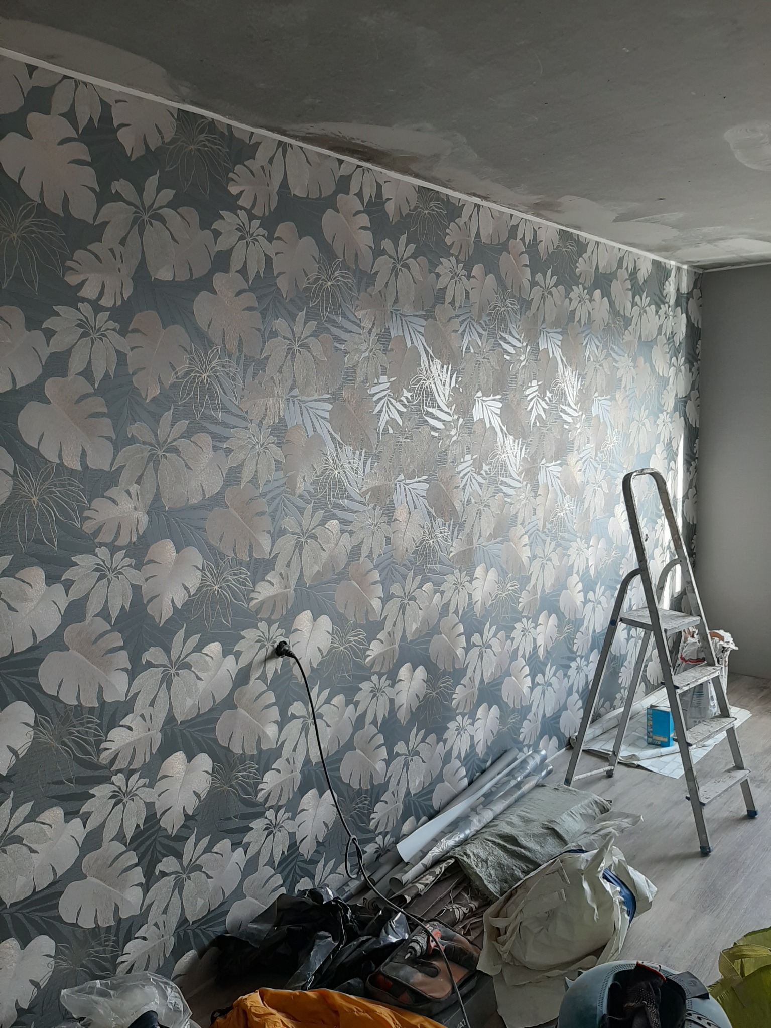 Ściana z nowo położoną tapetą w motywy roślinne, widoczna drabina malarska i narzędzia na podłodze, surowy sufit w tle.