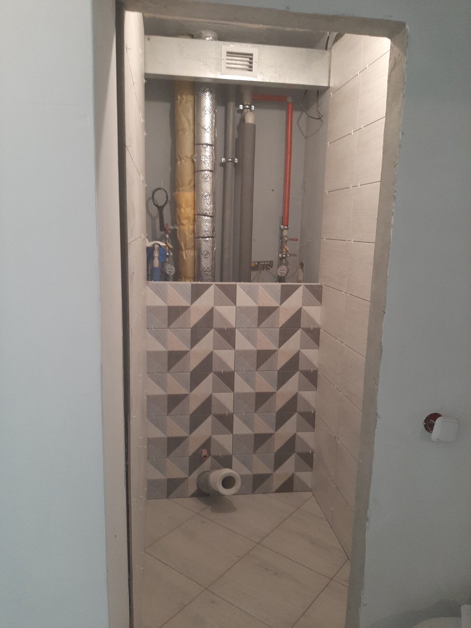 Fragment łazienki w trakcie prac: częściowo ułożona mozaika z płytek w odcieniach szarości, widoczne rury instalacyjne i otwór kanalizacyjny.