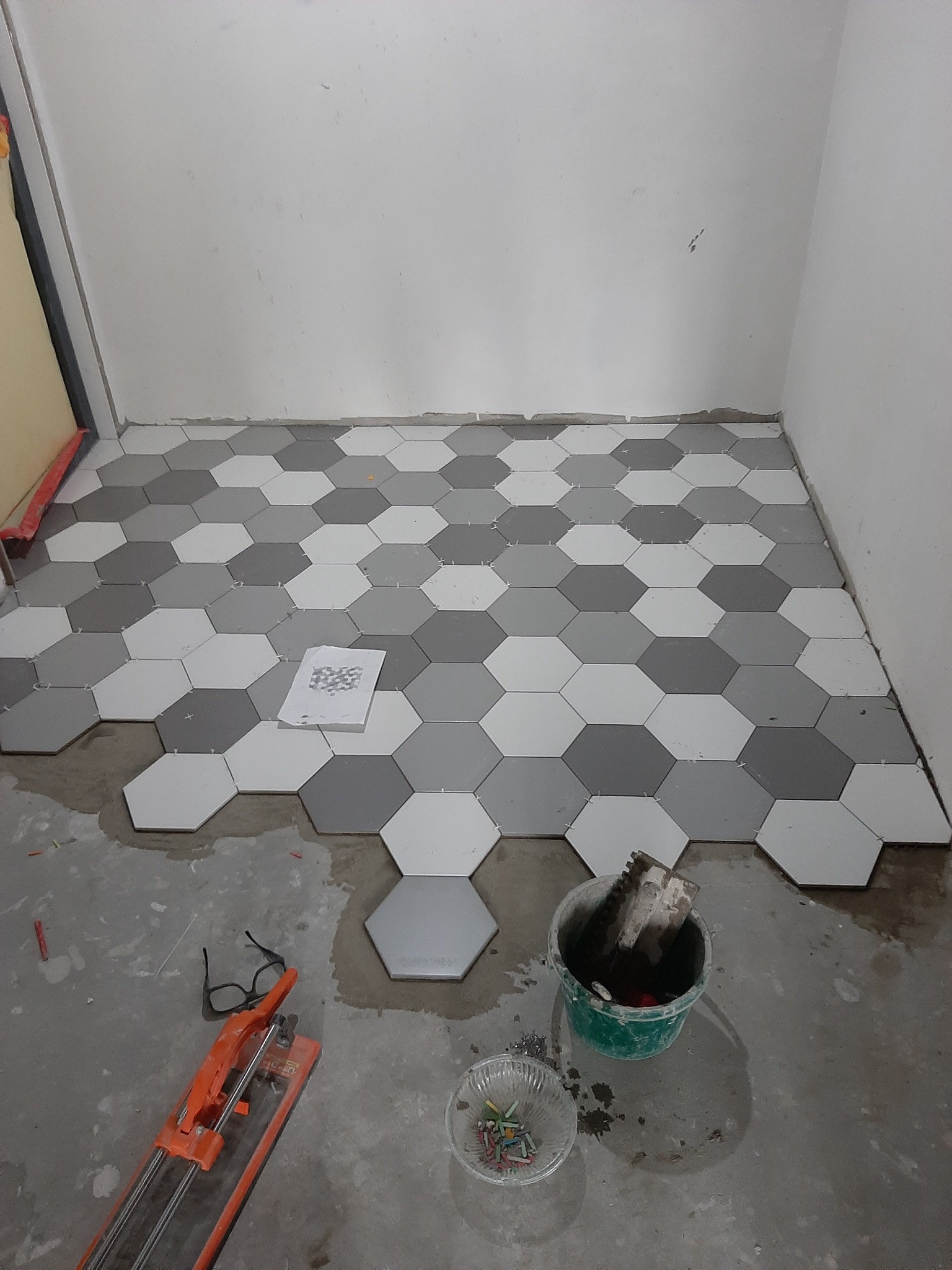 Układanie heksagonalnych płytek podłogowych w odcieniach szarości i bieli, widoczny plan ułożenia, narzędzia glazurnicze i okulary na posadzce.