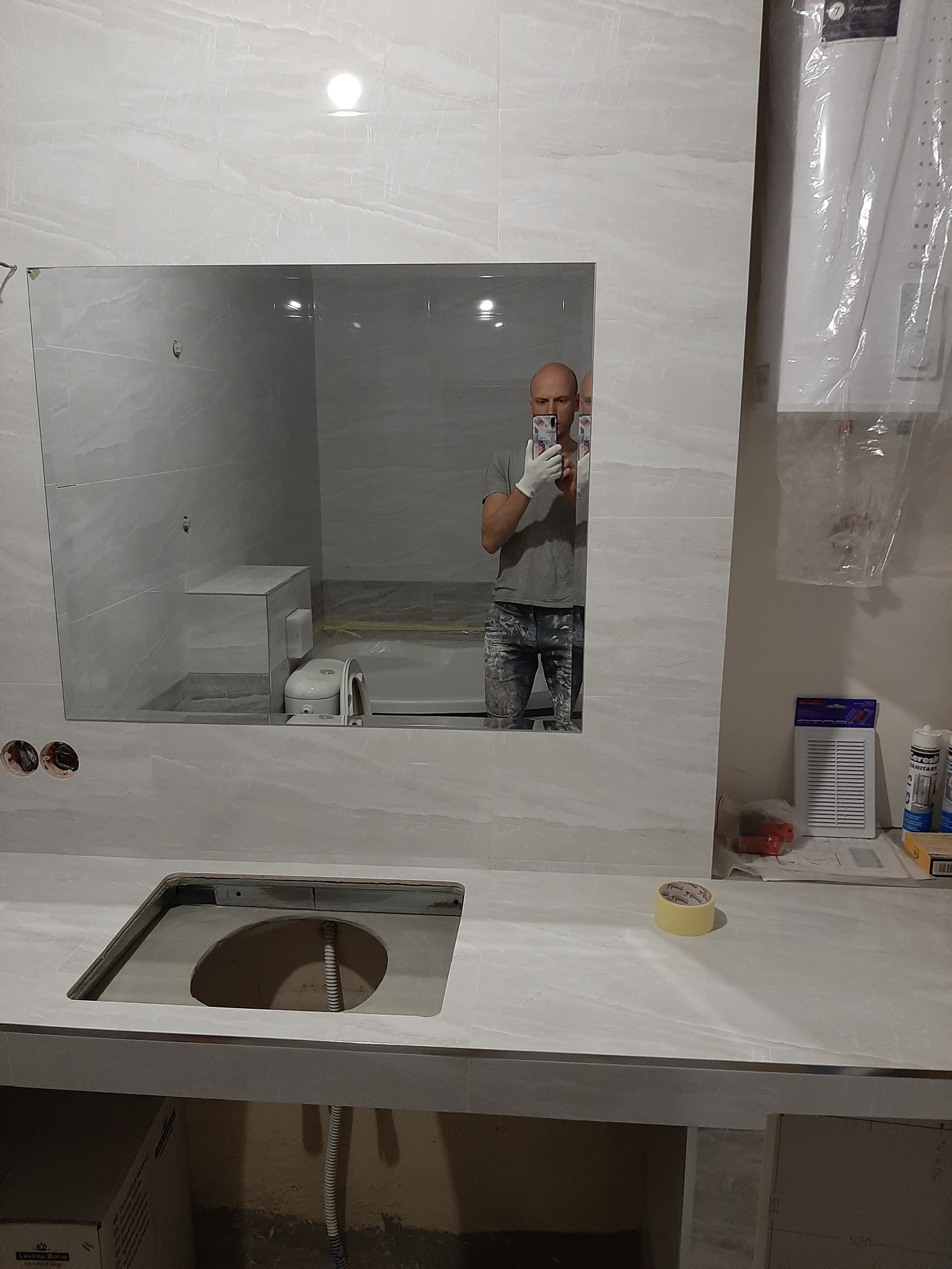 Niedokończona łazienka z białymi płytkami, widoczne lustro z odbiciem osoby robiącej zdjęcie, umywalka bez baterii i zabezpieczony folią bojler.