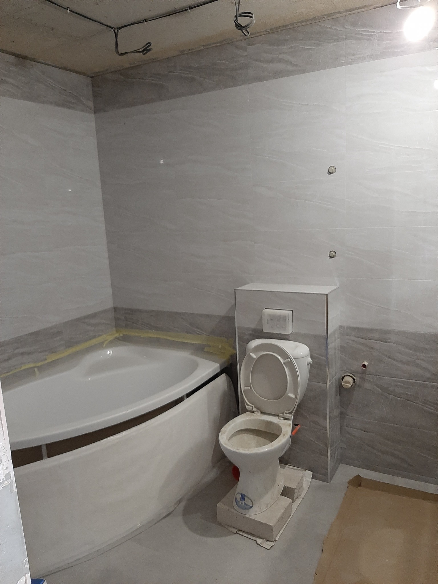 Wnętrze łazienki w trakcie prac: narożna wanna z zabezpieczeniem folią, podwieszana toaleta na podwyższeniu z betonu komórkowego, szare płytki imitujące kamień na ścianach.