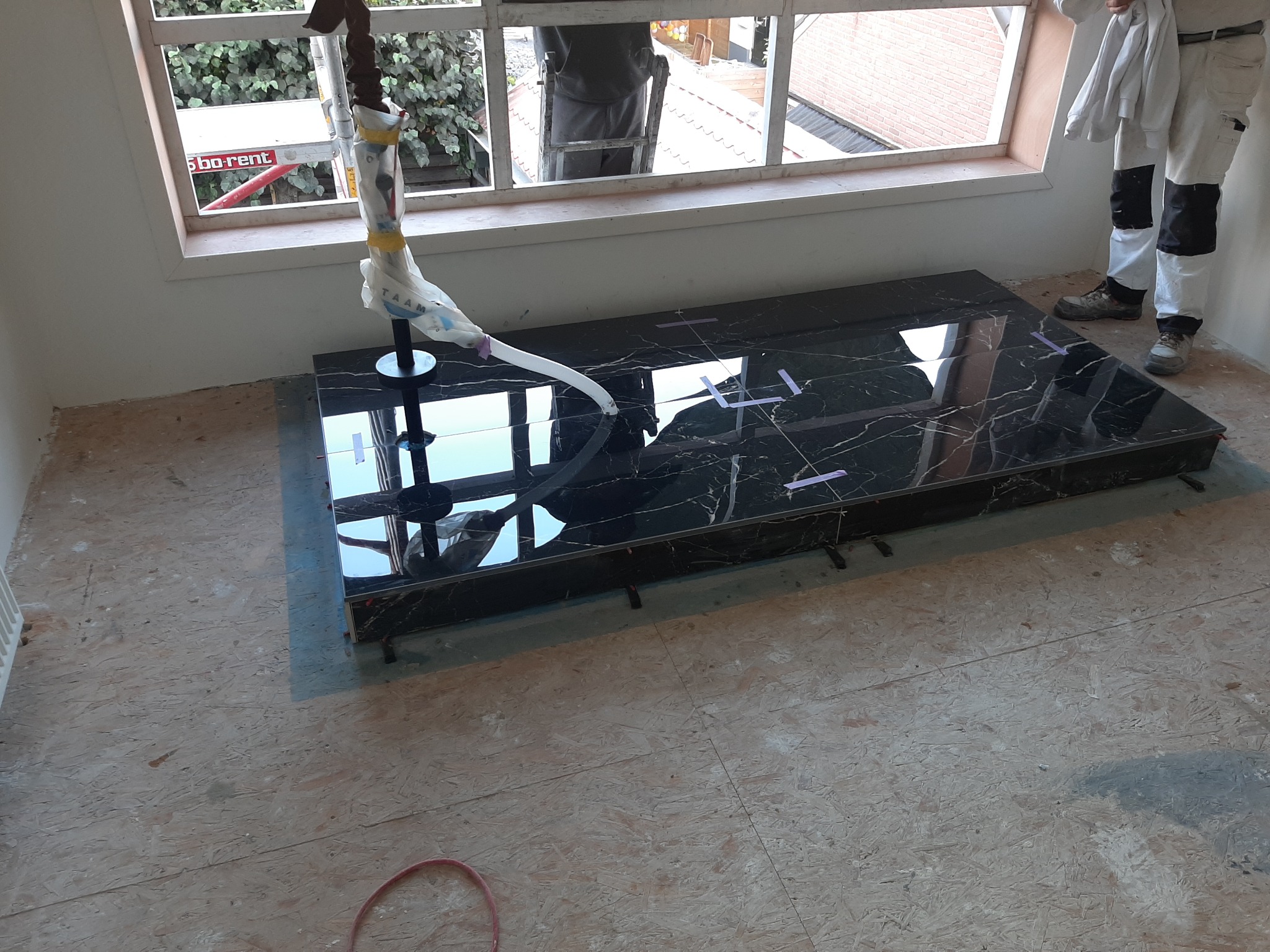 Montaż czarnego, błyszczącego blatu z marmurkowym wzorem w łazience, widoczne podkładki poziomujące, pracownik w ubraniu roboczym, okno z widokiem na zewnątrz, podłoga w trakcie prac wykończeniowych.