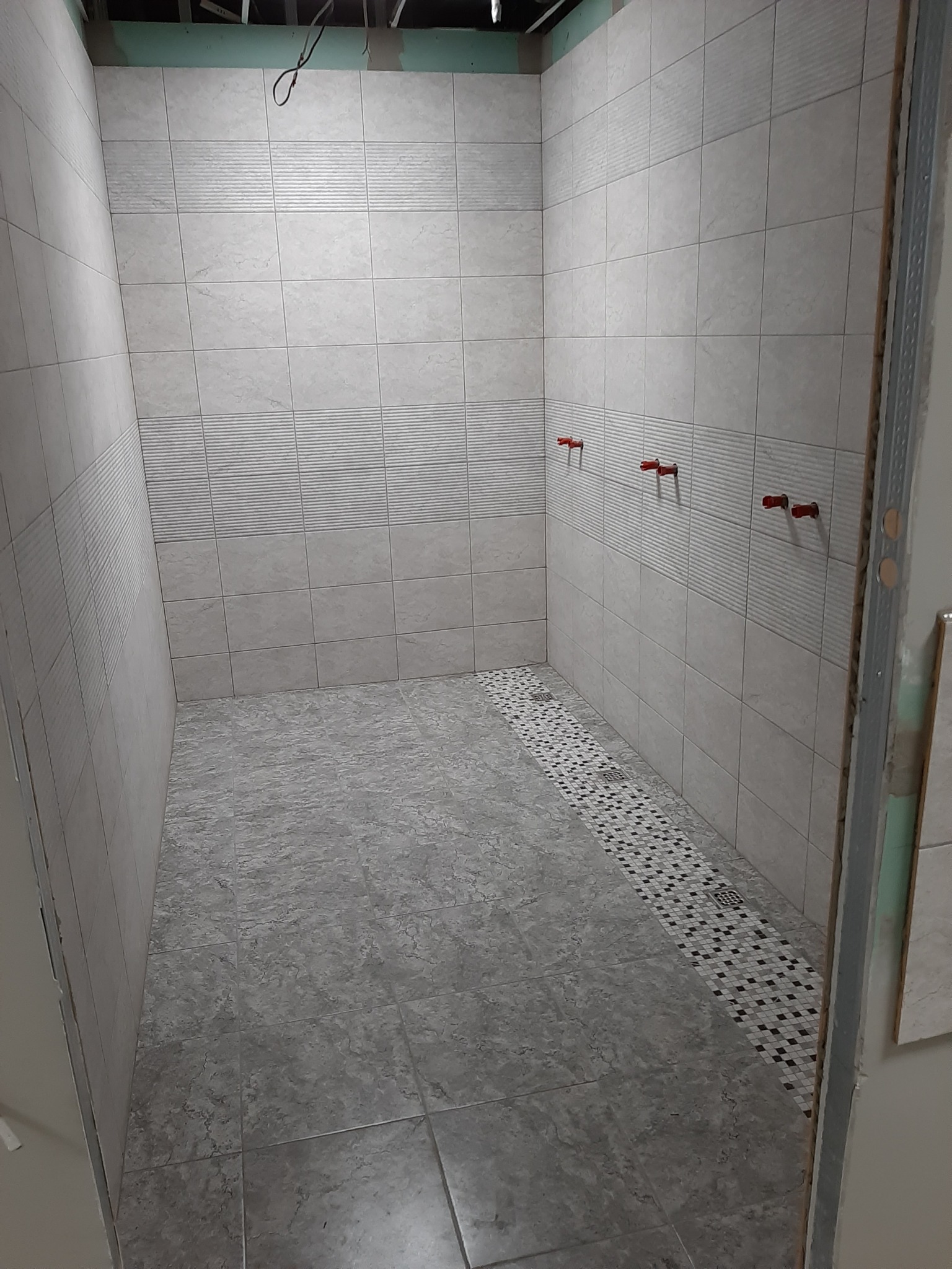 Wnętrze wykończonej kabiny prysznicowej z szarymi płytkami na ścianach i podłodze, widoczne przyłącza wodne z czerwonymi zaślepkami i odpływ liniowy wyłożony mozaiką.