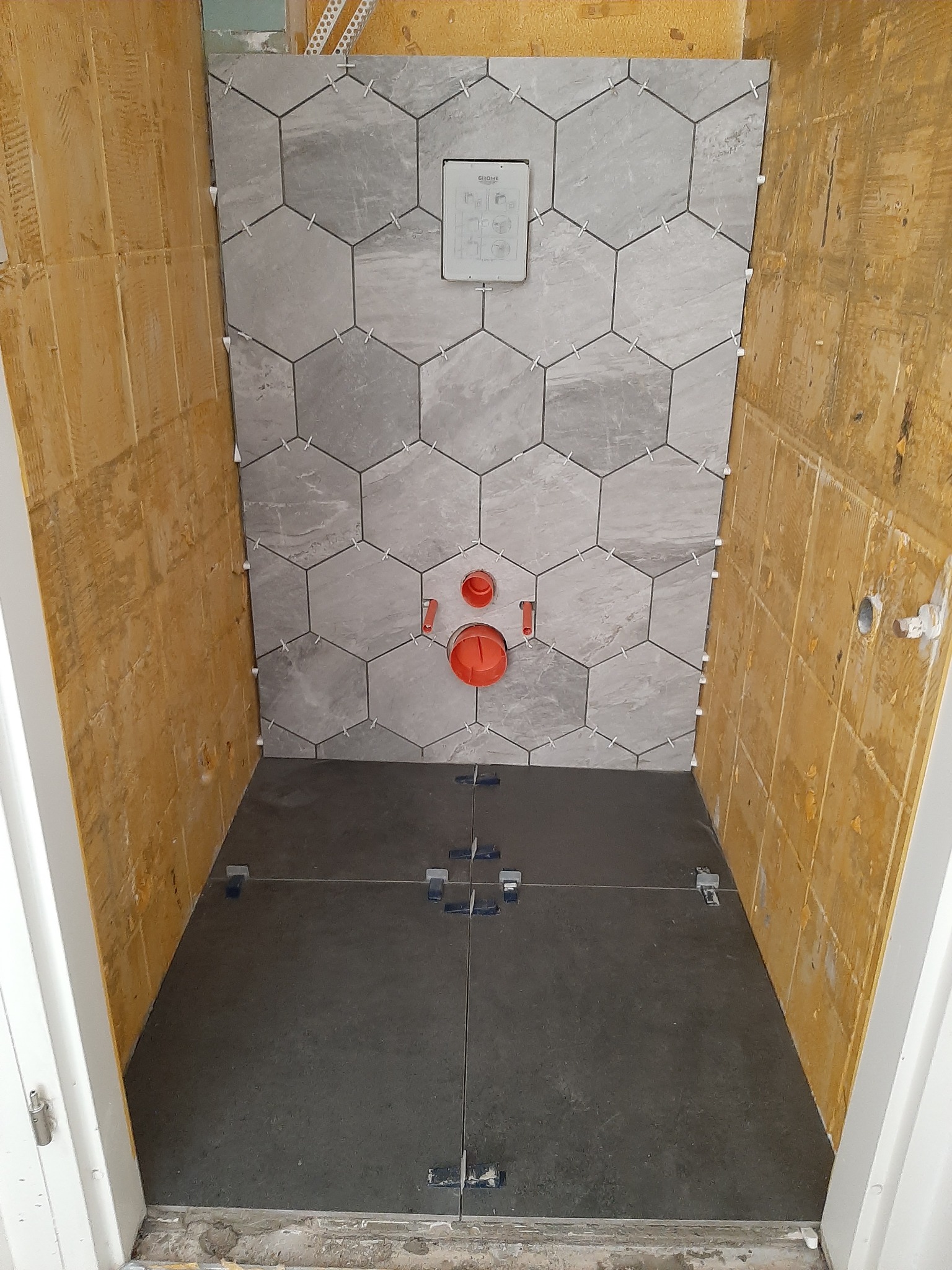 Montaż szarych, heksagonalnych płytek ściennych w łazience z widoczną instalacją podtynkową i pomarańczowymi elementami hydraulicznymi, płytki podłogowe w trakcie układania z użyciem krzyżyków...