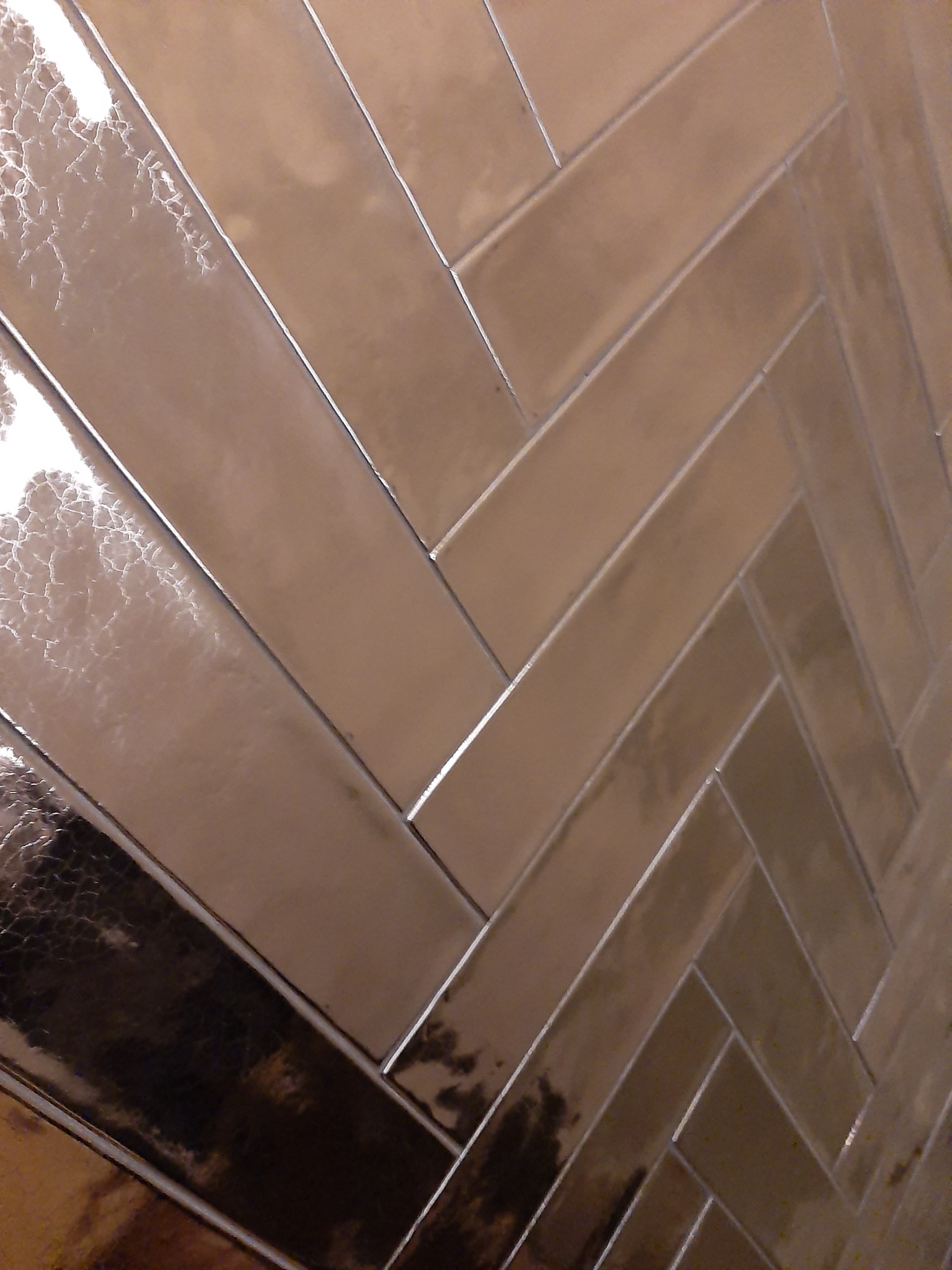 Połyskujące, beżowe płytki ułożone w jodełkę, z widocznymi refleksami światła i drobnymi niedoskonałościami powierzchni.