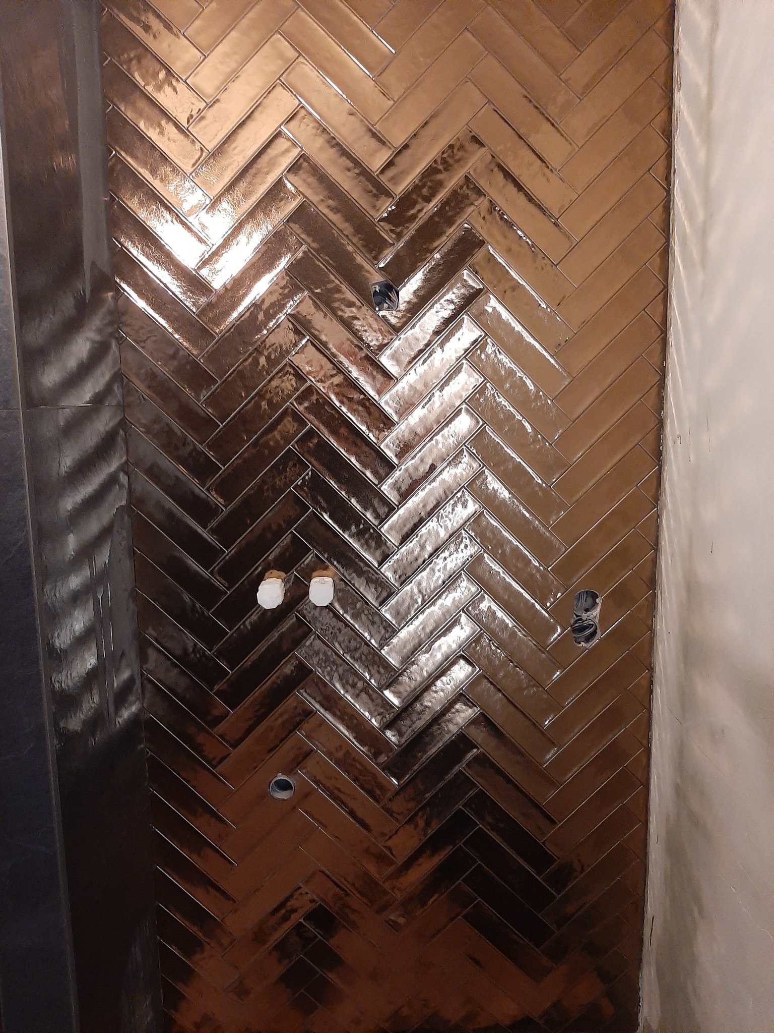 Błyszczące, brązowe płytki ułożone w jodełkę na ścianie, widoczne otwory na instalacje elektryczne i fragment surowej ściany.