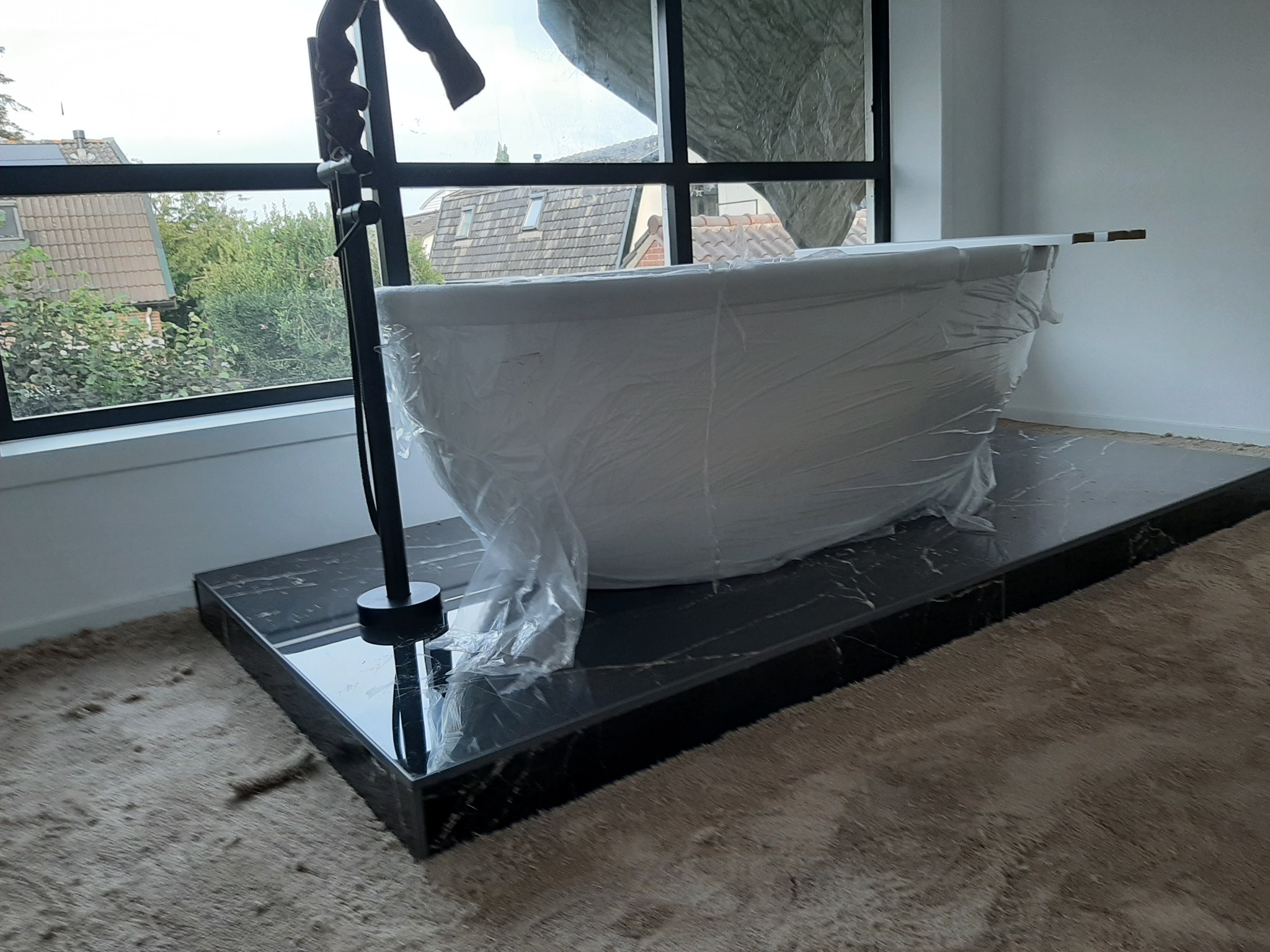 Nowa wanna owinięta folią ochronną, czarna bateria wolnostojąca, marmurowa podstawa, duże okno z widokiem na domy i zieleń na zewnątrz.