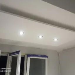 Sufit podwieszany z trzema punktowymi lampami LED, widoczne okna zabezpieczone folią malarską, ściany przygotowane do malowania.