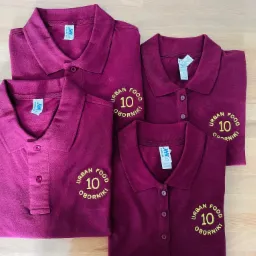 Cztery bordowe koszulki polo z haftowanym logo 'URBAN FOOD 10 OBORNIKI' ułożone na drewnianym blacie.