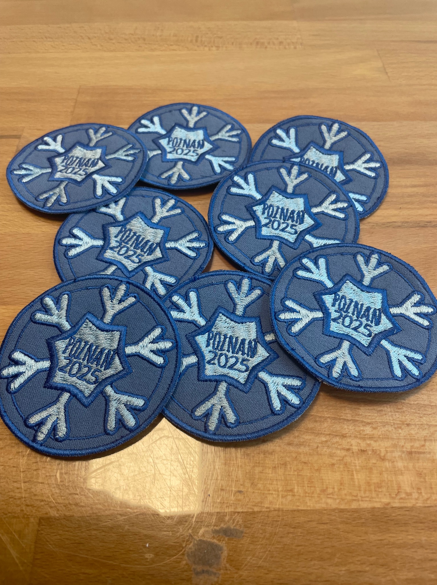 Stos okrągłych naszywek z motywem śnieżynki i napisem 'Poznań 2025' na drewnianym stole, wykonanych techniką haftu maszynowego, w odcieniach błękitu i szarości.